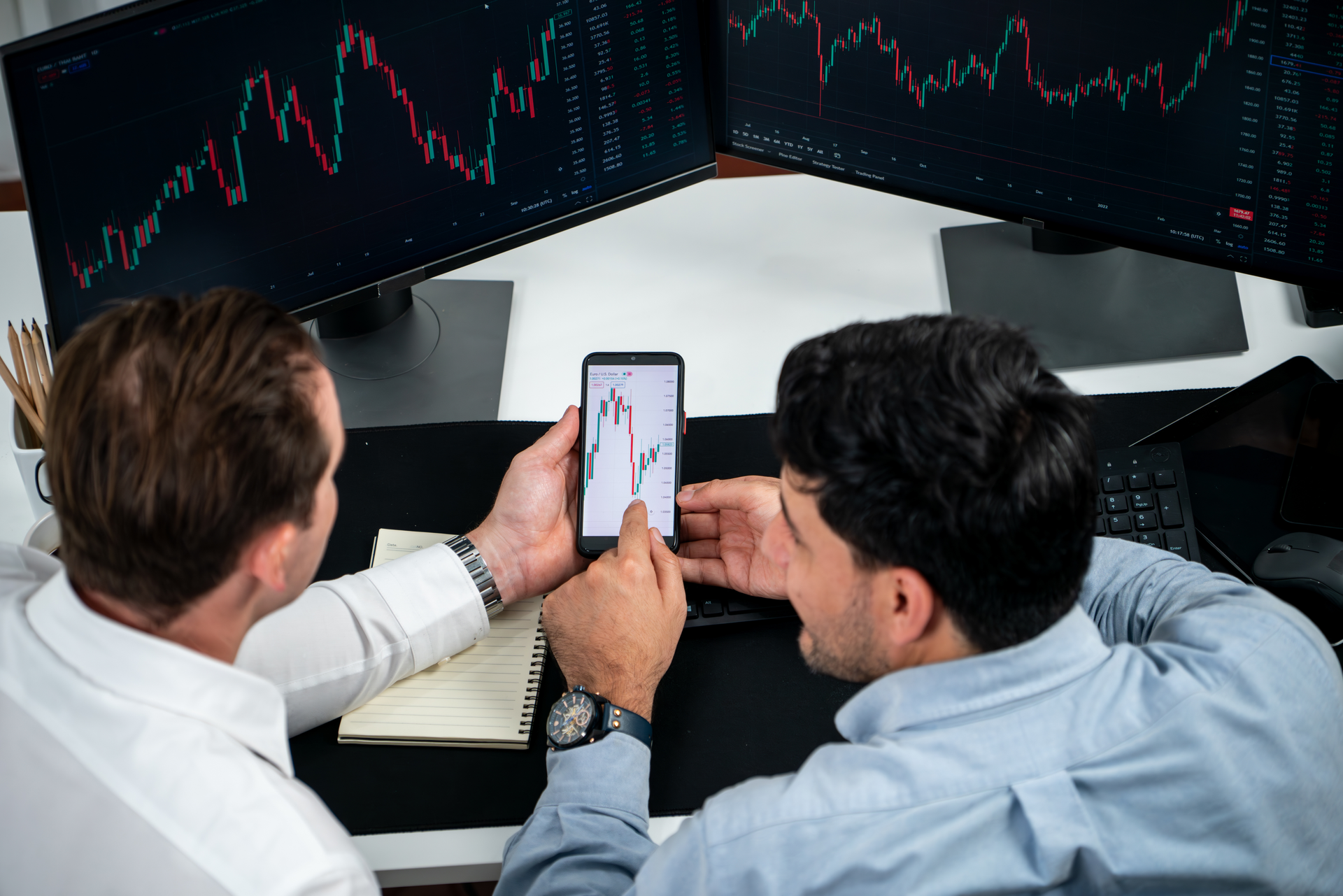 Two business professionals analyze stock market charts on multiple screens, discussing financial investments and market trends.