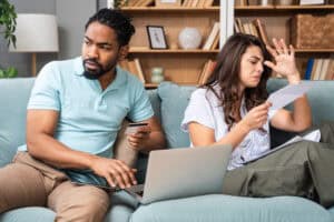 A concerned couple sitting on a couch, reviewing bills and using a laptop, symbolizing financial stress and mortgage payment challenges.