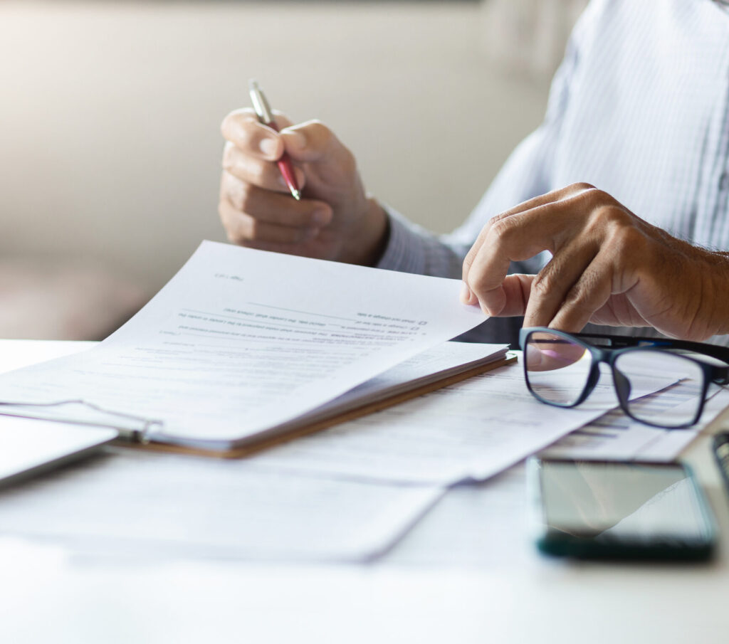 Person reviewing loan estimate paperwork with pen in hand, glasses and laptop on desk